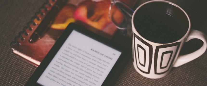 Photo of eReader, glasses, pen, notebook and mug of coffee