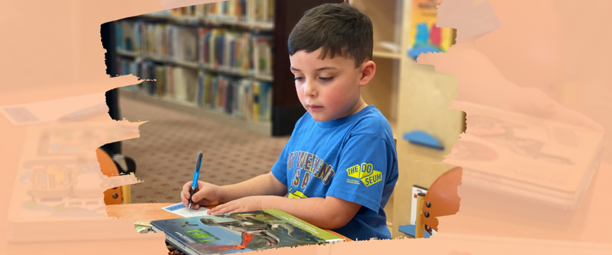 Boy sitting at library table looking at book and writing on Kids Pick bookmark