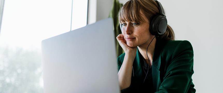 Woman wearing headphones and watching a computer screen. A window and plant are in the background.