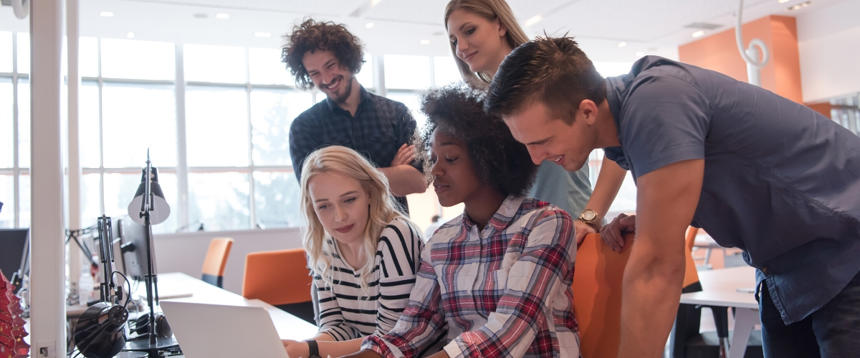 Two women and two men looking over the shoulder of a woman as she works on a laptop.