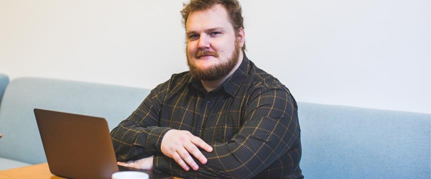 Man sitting at table with arms crossed and laptop and coffee mug.