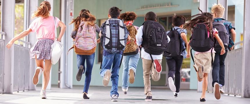 A group of eight elementary school students wearing back packs running into the school building.