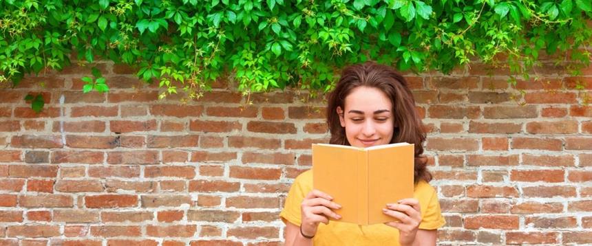 Woman reading a book in front of a brick wall with plants 
