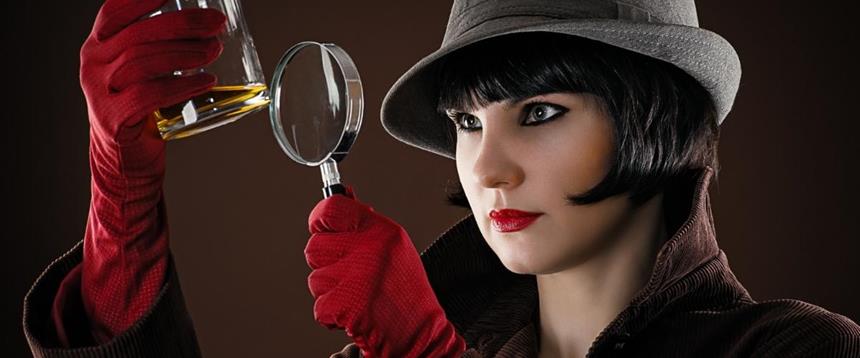 Woman wearing hat a gloves holding up a magnifying glass to a glass with liquid inside