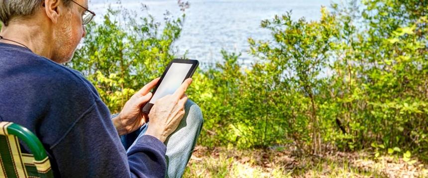 Man sitting in a lawn chair on the bank of a river or lake using an e-Reader.